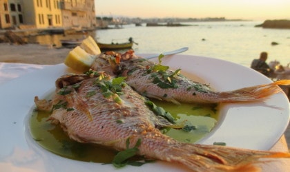 Whole baked fish on plate with olive oil at a restaurant, sea and old buildings in the background