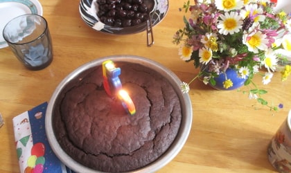 a chocolate cake in a round metal pan with two candles burning on the cake and a bouquet of flowers and a bowl of cherries near it