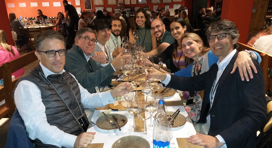 people at a long table in a winery, happily proposing a toast with lifted wineglasses